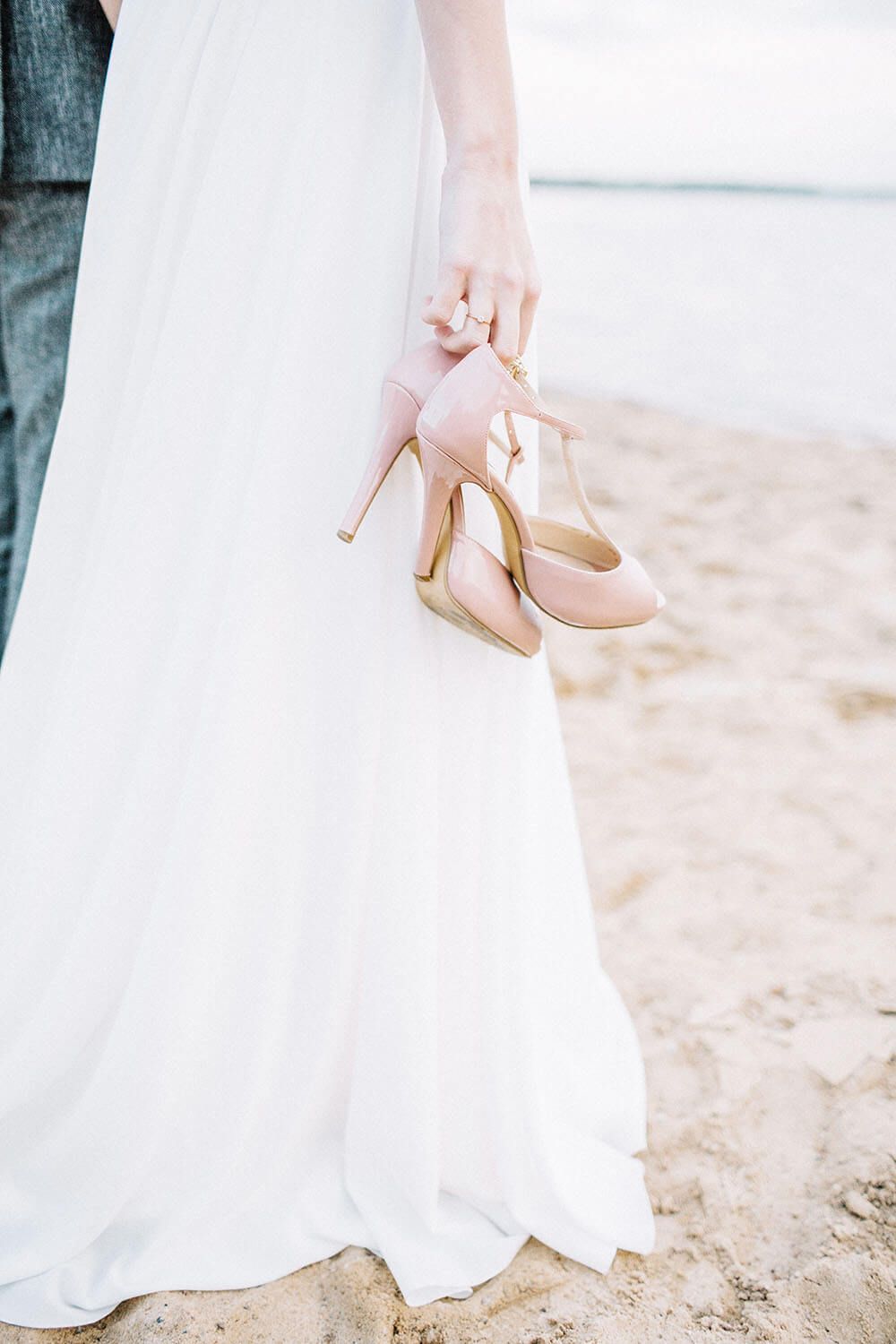 Bride holding pink high heels at the beach, wedding shoes for special occasion.