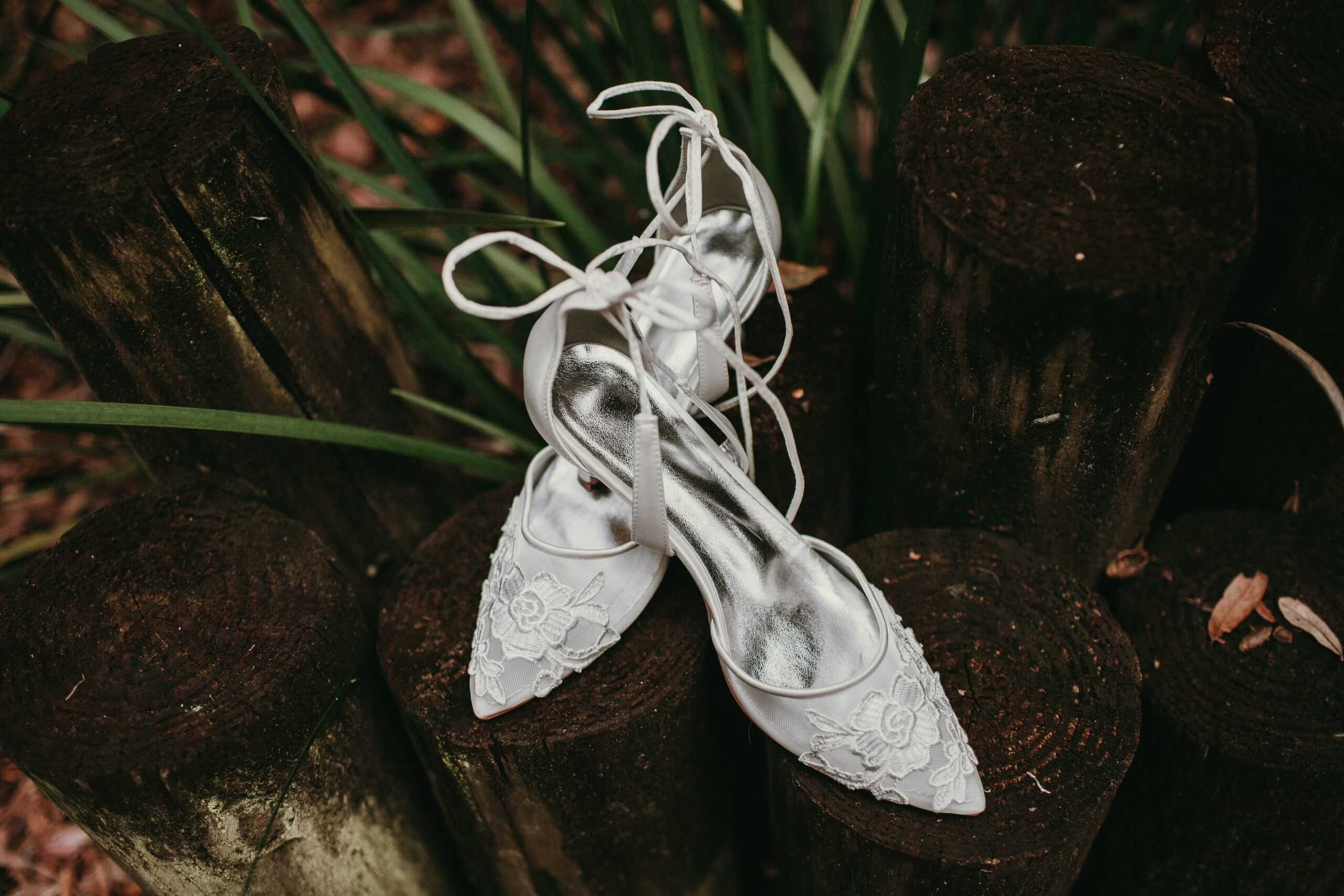 Elegant white lace wedding shoes with ankle straps on rustic wooden logs.