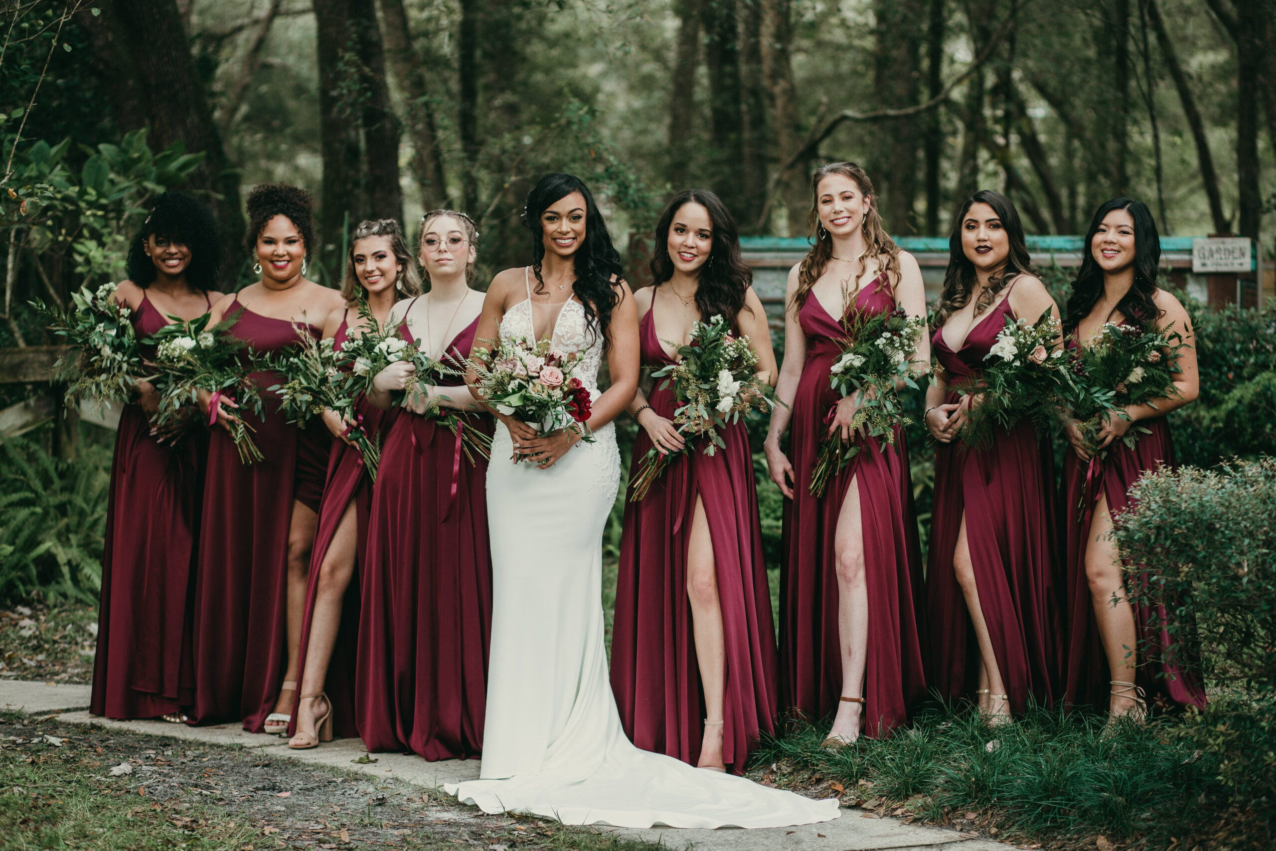 Bridal party with bride and bridesmaids in burgundy dresses holding bouquets outdoors.