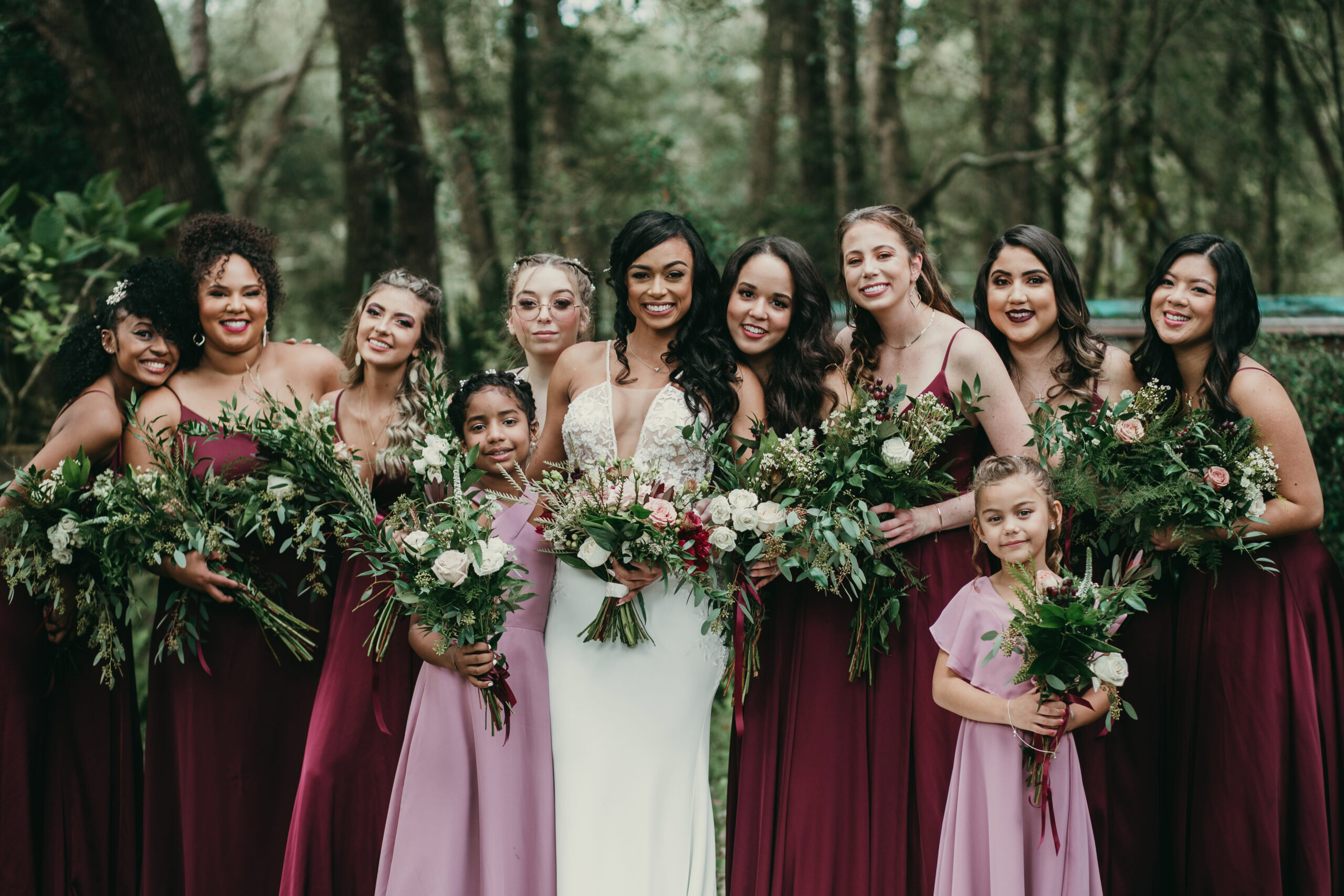 Bridesmaids and bride in a forest, holding bouquets, celebrating a wedding outdoors.