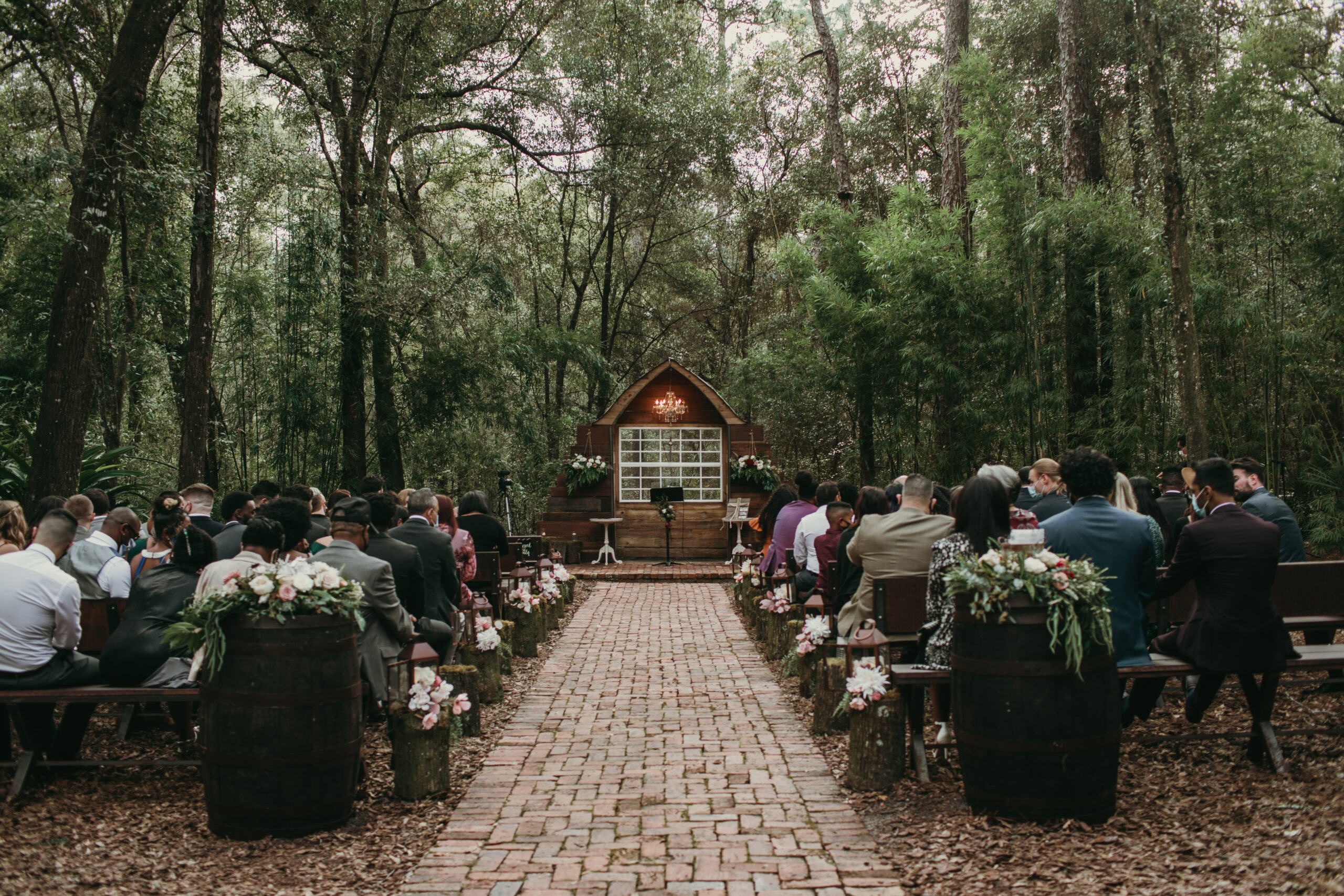 Wedding ceremony in a forest with guests seated on benches, surrounded by tall trees and lush greene.