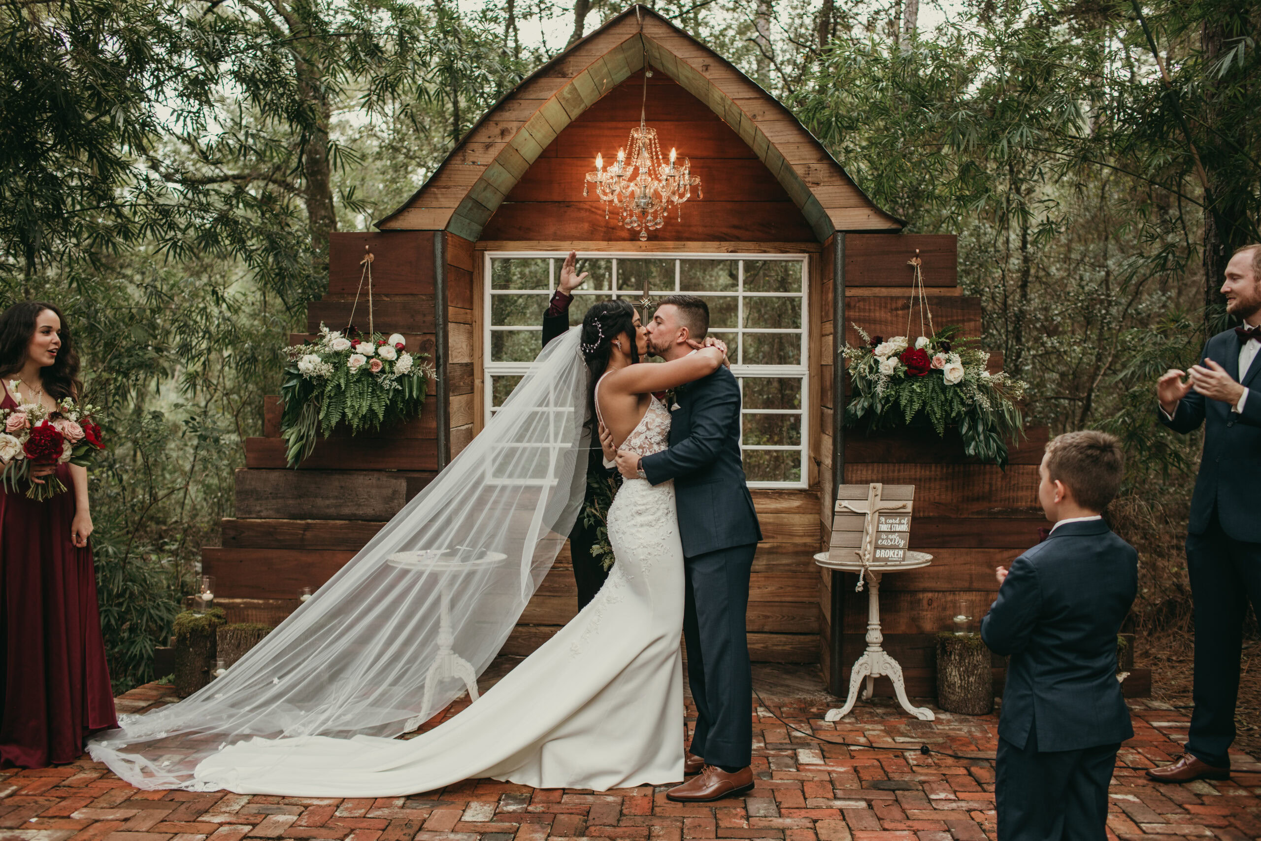 Wedding kiss at outdoor ceremony with rustic backdrop and chandelier.