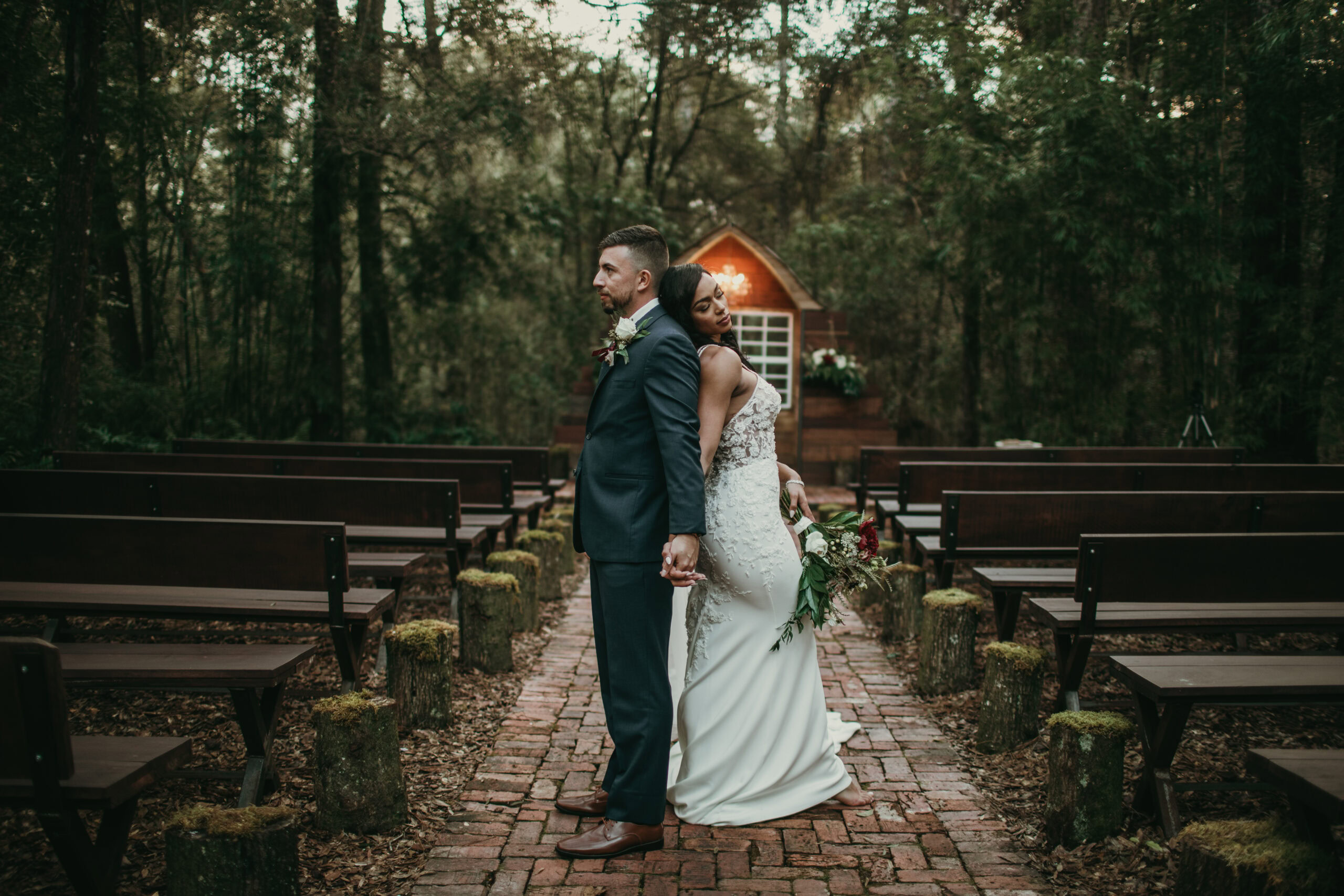 A couple stands back-to-back on a brick path in a forested outdoor wedding venue.