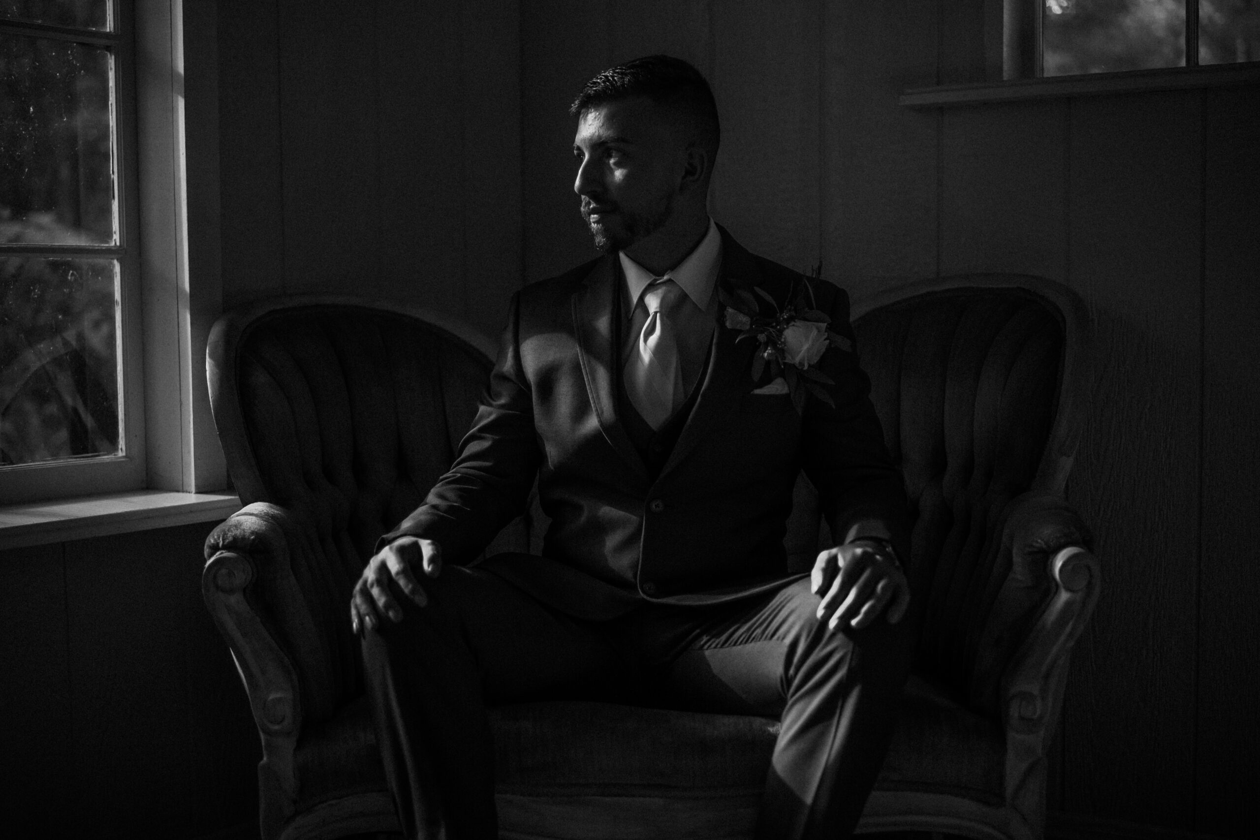 Groom in a suit sitting on a vintage sofa, preparing for his wedding day, with natural light coming.
